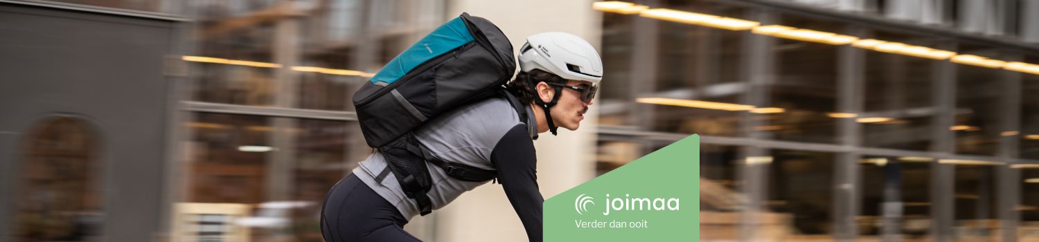 A cyclist wearing a helmet and carrying a Joimaa backpack rides swiftly past a building.
