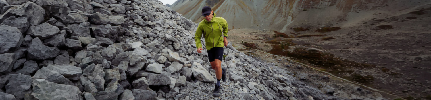A person in a green jacket and black shorts running on a rocky mountain trail.