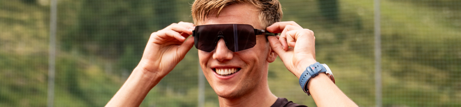 A person smiling while adjusting black sports sunglasses outdoors.