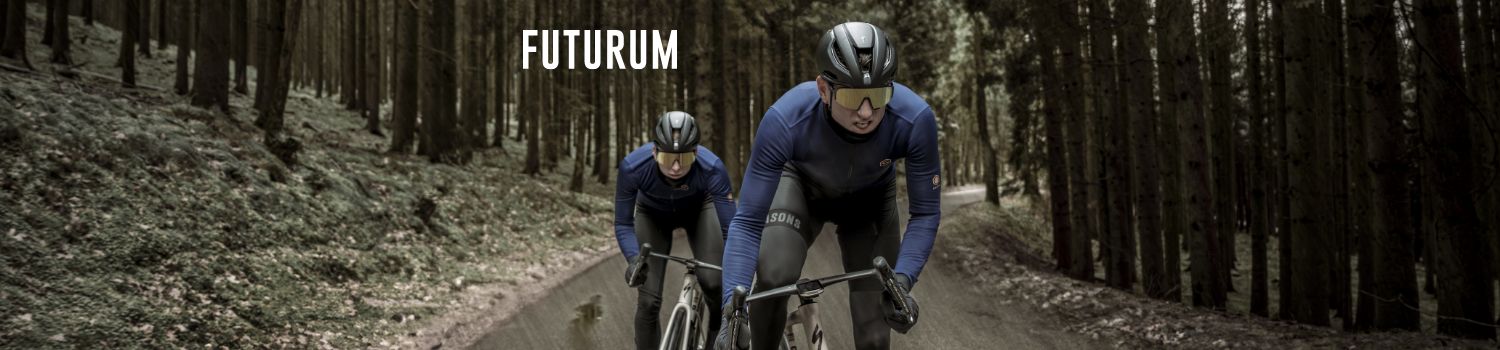 Two cyclists wearing blue jerseys and helmets ride through a forest path. The word 'Futurum' is visible in the background.