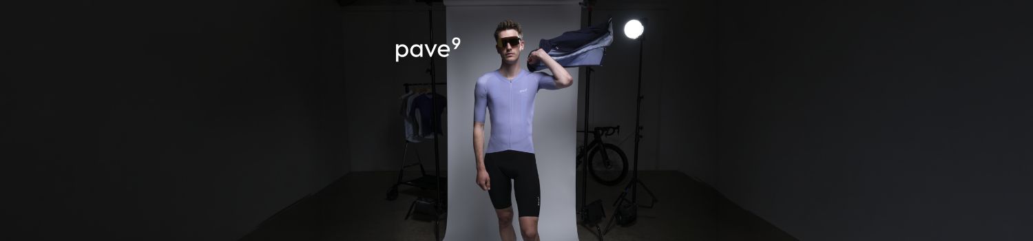 A cyclist wearing a light purple Pave9 jersey and black shorts poses in a studio setting with a bicycle in the background.
