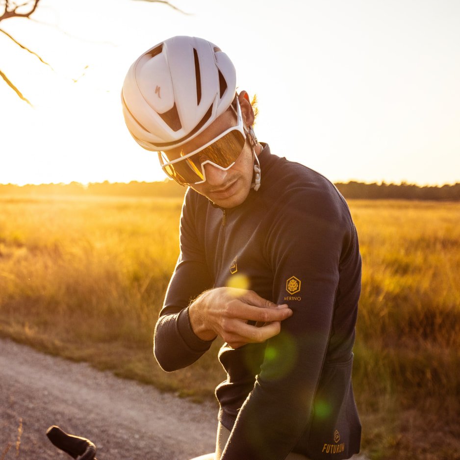 A cyclist wearing a white helmet and sunglasses points to a Futurum Merino jersey in a sunny outdoor setting.
