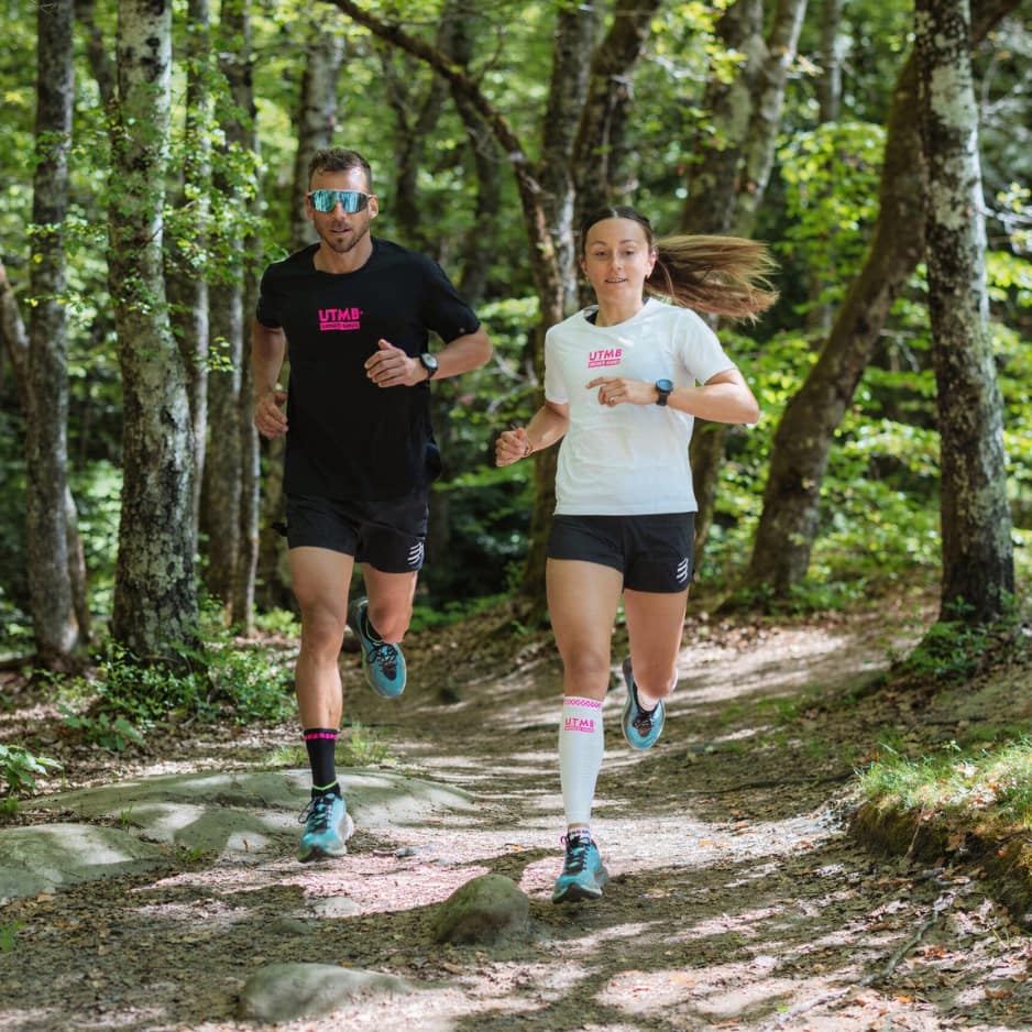A man and a woman running on a forest trail wearing UTMB branded shirts and shorts.