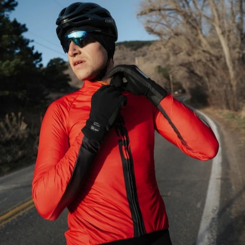 A cyclist wearing a red jacket and black helmet is adjusting his zipper on a road.